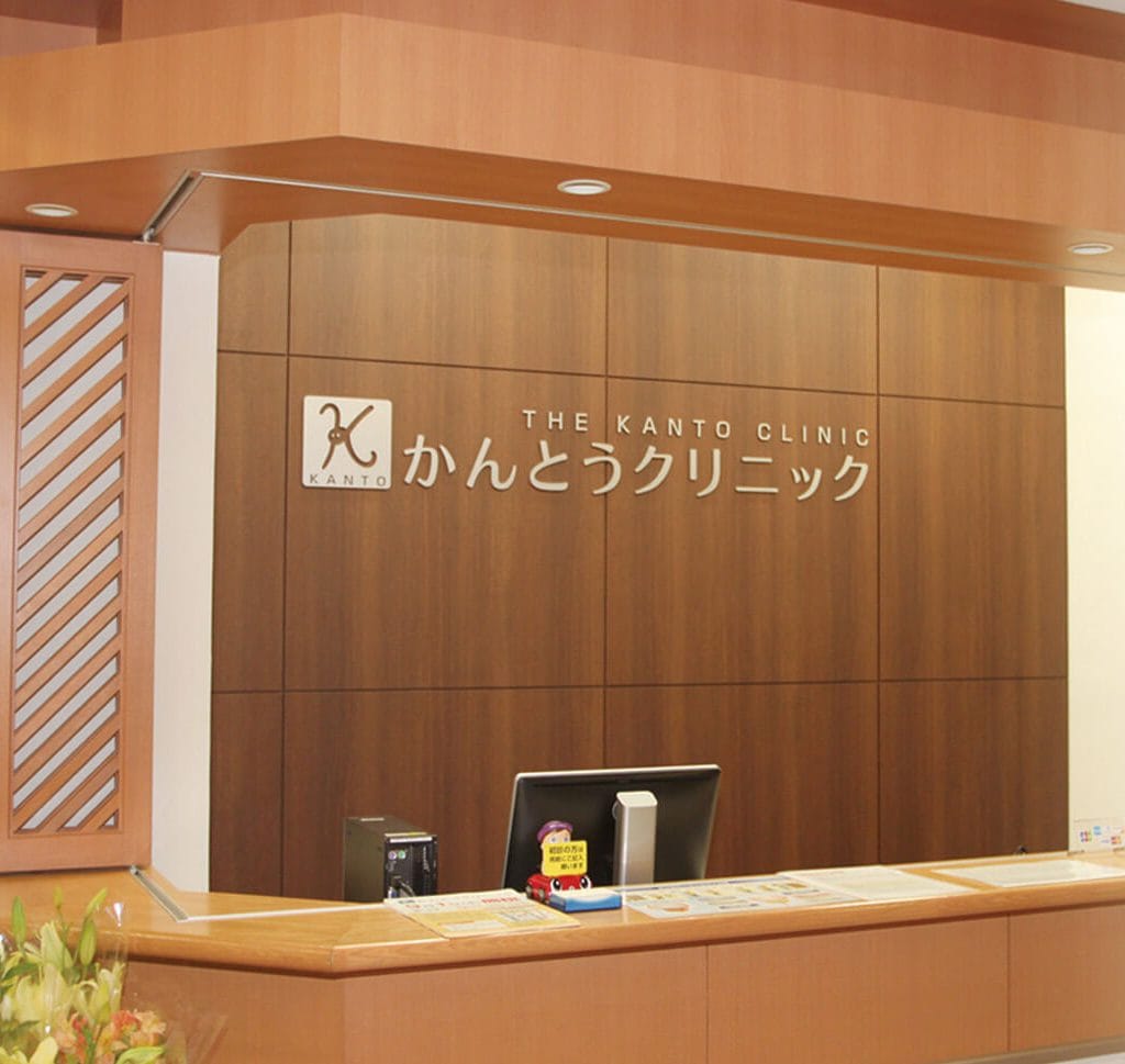 Reception desk at Kanto Clinic. The desk is a light-colored wood, with a plant next it and the receptionist's computer can be seen beyond the reception counter. The back wall is a darker wood, and silver metal letter outline the Kanto Clinic logo on the back wall.